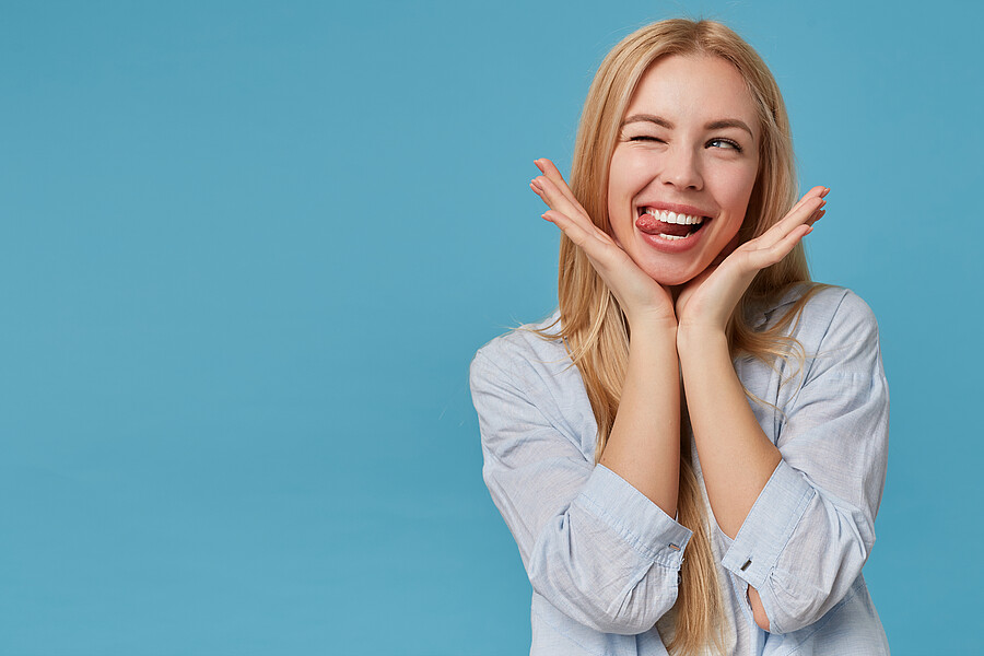 Portrait of pretty young blonde lady with long hair keeping head on raised hands, making faces over blue background, looking aside joyfully with closed eye and showinng tongue